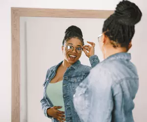 A woman in a denim jacket and glasses looks at her reflection in the mirror looking happy