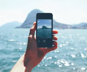 A person holding their phone in front of a scenic landscape of the sea and an island as they are about to take a photo