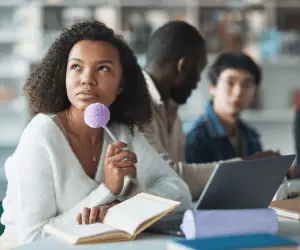 2 males in the background with a girl up close holding a purple fluffy pen lent on her chin in deep thought