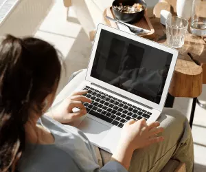 An overhead shot of a woman with brown hair sat on a chair in her home typing on her laptop