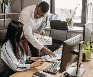 A man and woman collaborate on a computer in a modern office setting, focused on their work