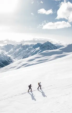 2 skiers walking up a snowy hill wearing their skis on a sunny day