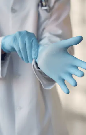 A person wearing a white coat and putting on blue gloves, standing in a clinical or laboratory setting