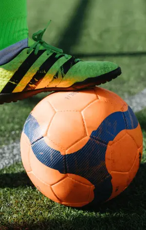 A close up of a person's foot wearing a football boot resting it on top of an orange football on a football pitch