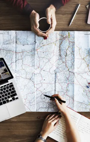 Two people sat at a table with a map open with a laptop open too as they discuss destinations on the map