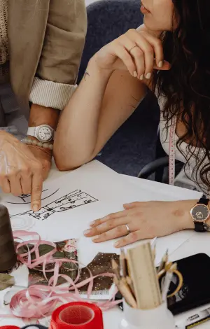 Two females reviewing a clothing design on a piece of paper surrounded by fabrics and threads
