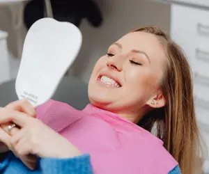 A woman sits in a dentist chair, holding a mirror as she looks at her teeth
