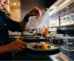 A chef wearing a black apron serving up a meal and sprinkling some extra seasoning on top