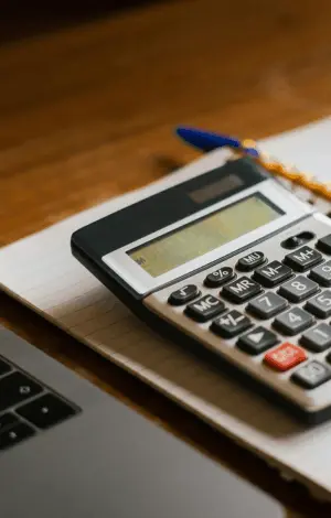 A close up of a calculator sat on top of a notebook next to a laptop on a desk