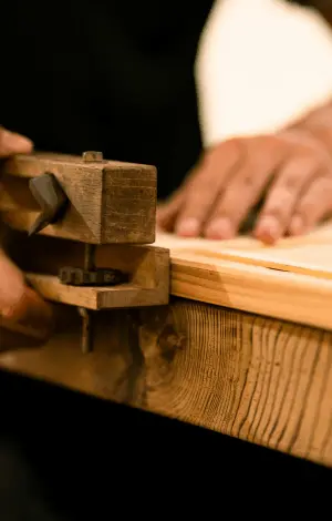 A close up of a guy holding a piece of wood on a table and cutting it with a wooden tool